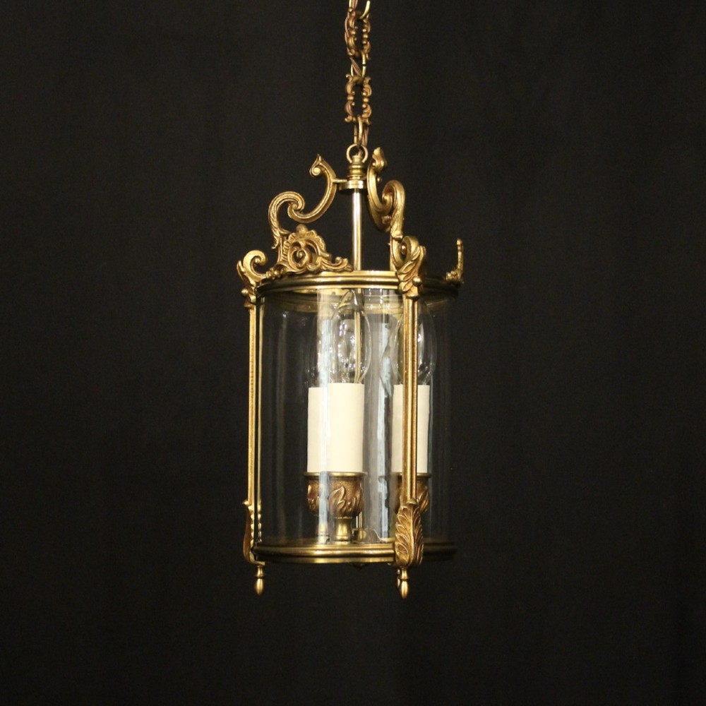 An antique French gilded bronze hall lantern featuring a cylindrical glass shade, three central candle sleeves, and ornate cast scrollwork mounts suspended from a chain.