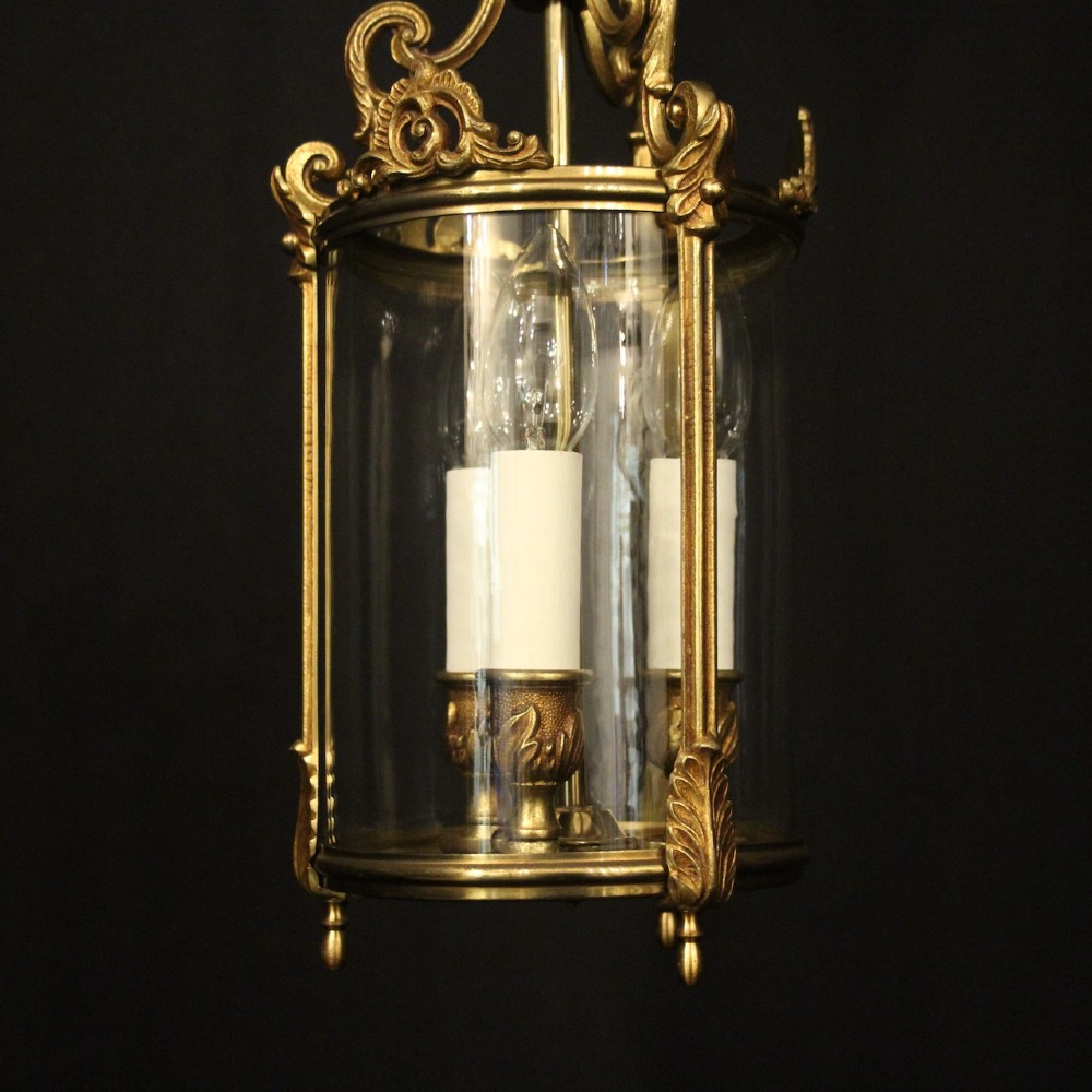 An antique French gilded bronze hall lantern featuring a cylindrical glass shade, three central candle sleeves, and ornate cast scrollwork mounts suspended from a chain. View II