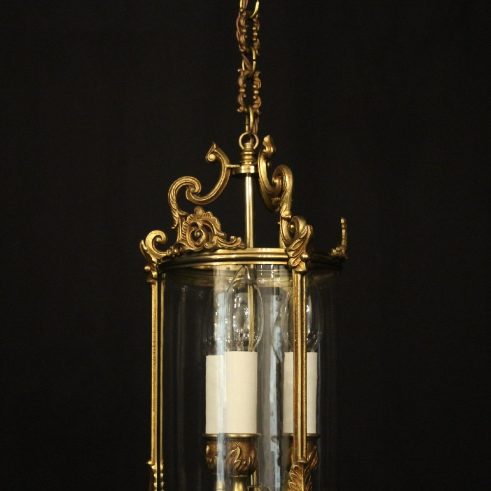 An antique French gilded bronze hall lantern featuring a cylindrical glass shade, three central candle sleeves, and ornate cast scrollwork mounts suspended from a chain. View III