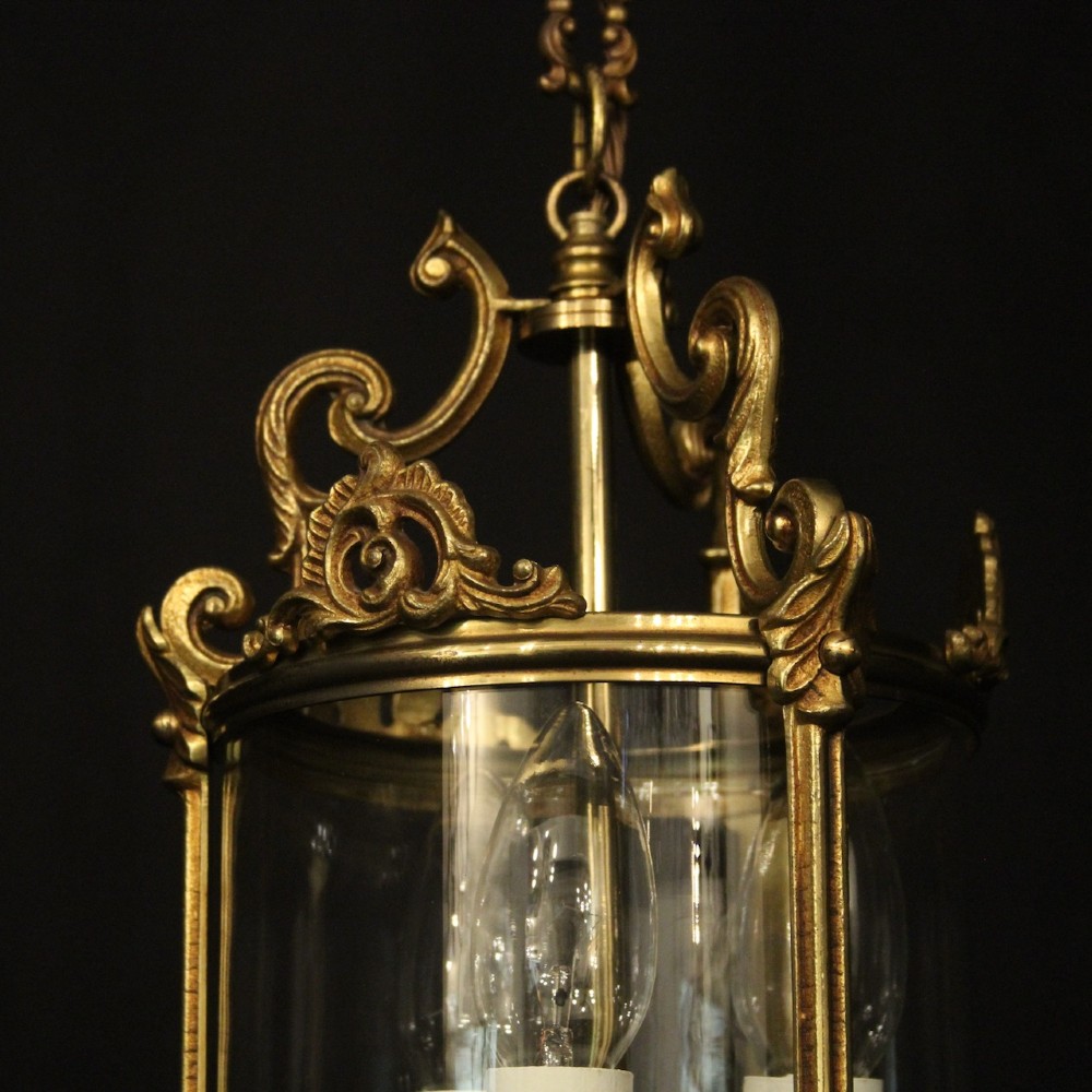 An antique French gilded bronze hall lantern featuring a cylindrical glass shade, three central candle sleeves, and ornate cast scrollwork mounts suspended from a chain. View IV