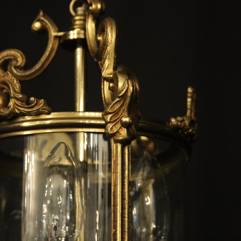An antique French gilded bronze hall lantern featuring a cylindrical glass shade, three central candle sleeves, and ornate cast scrollwork mounts suspended from a chain. View V