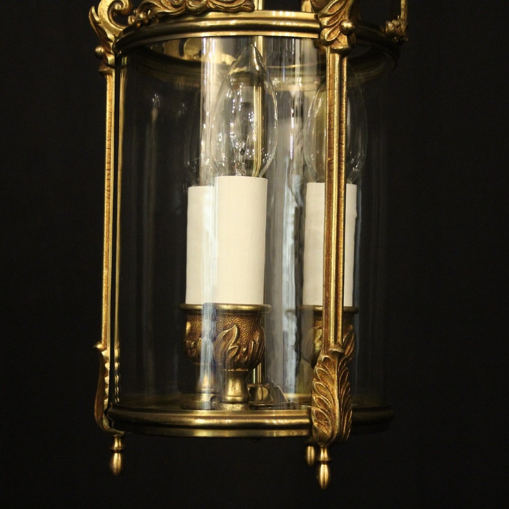 An antique French gilded bronze hall lantern featuring a cylindrical glass shade, three central candle sleeves, and ornate cast scrollwork mounts suspended from a chain. View VI