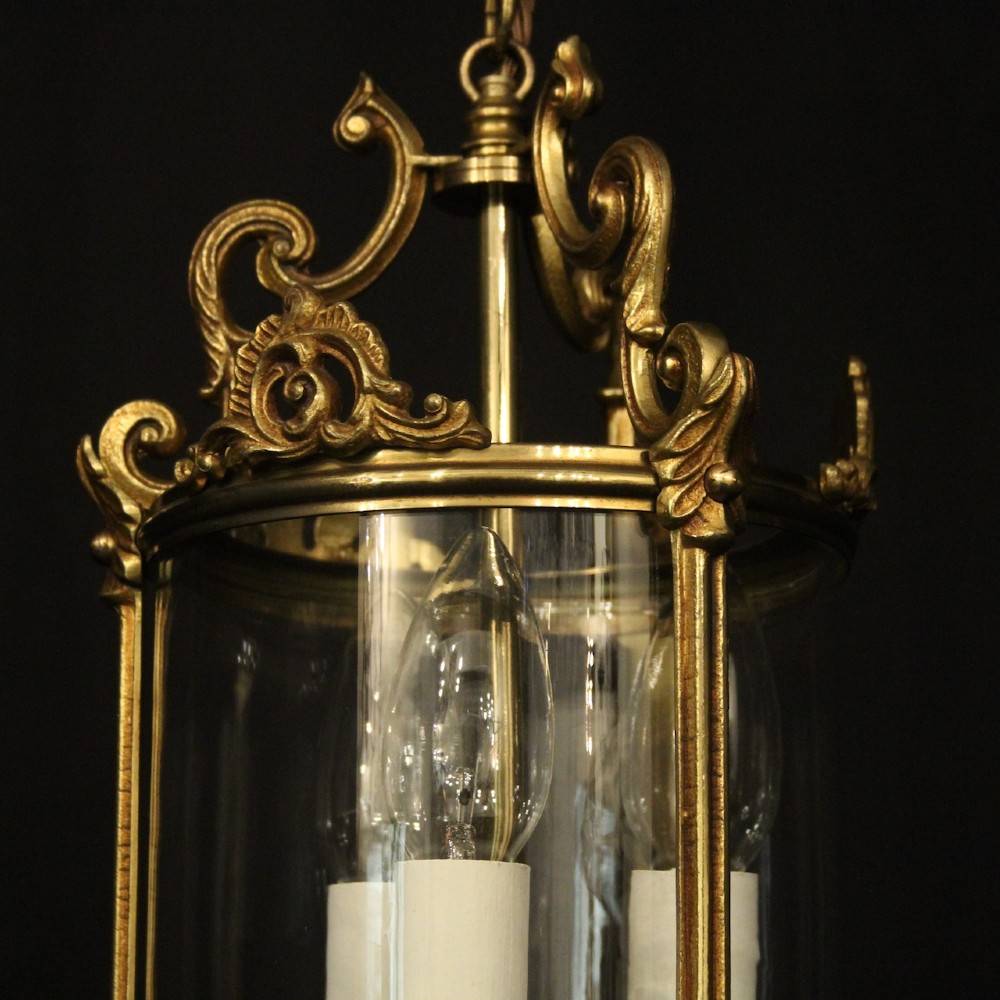 An antique French gilded bronze hall lantern featuring a cylindrical glass shade, three central candle sleeves, and ornate cast scrollwork mounts suspended from a chain. View VIII