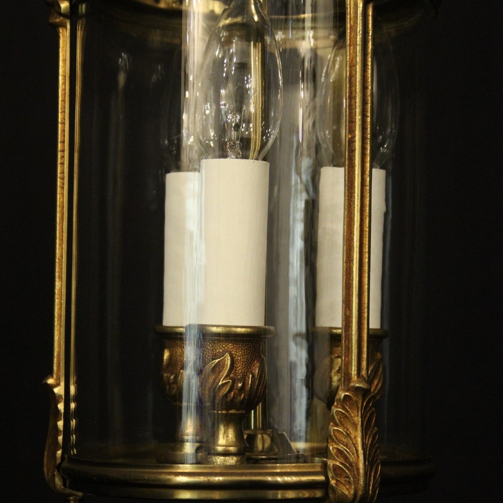 An antique French gilded bronze hall lantern featuring a cylindrical glass shade, three central candle sleeves, and ornate cast scrollwork mounts suspended from a chain. View IX