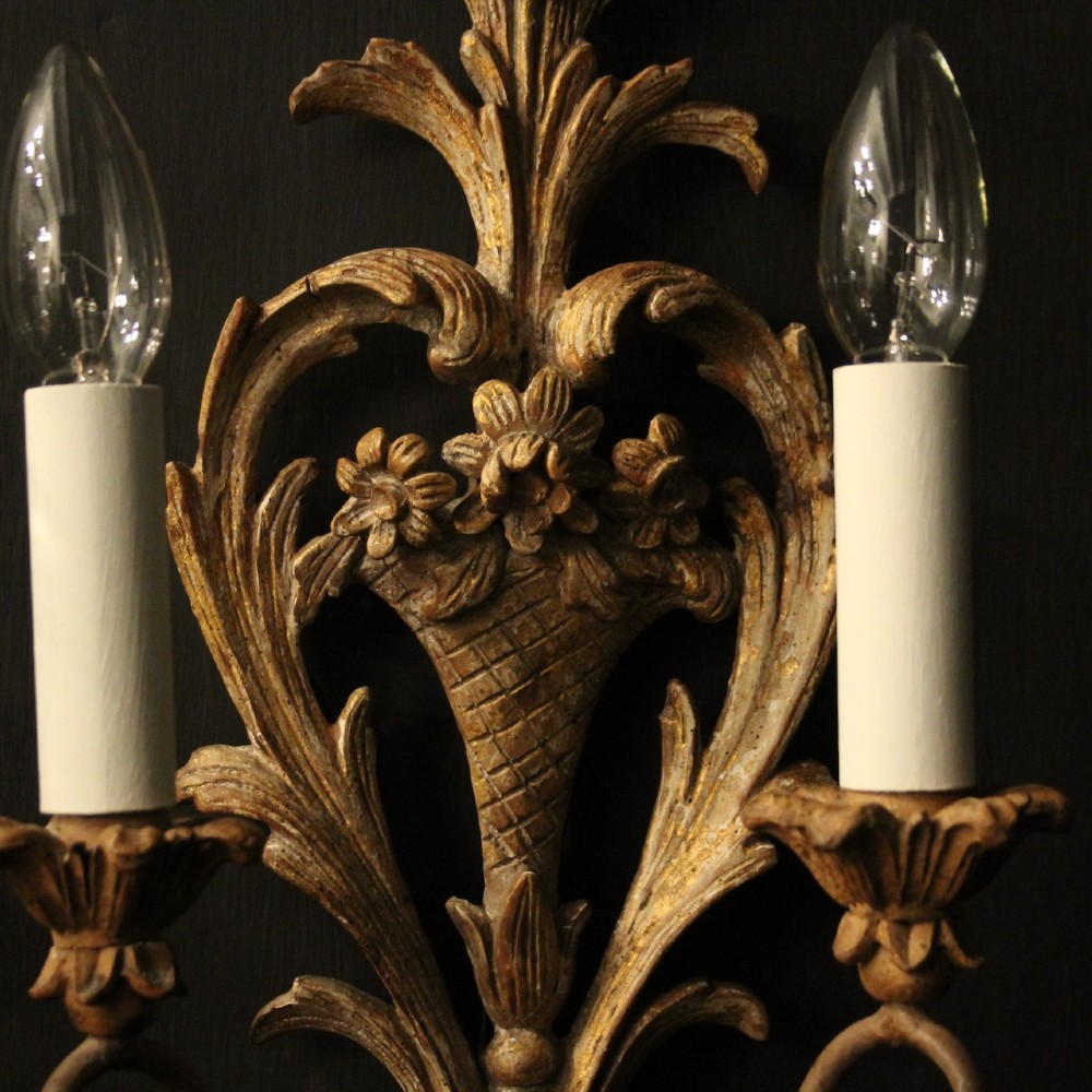 A pair of French carved wooden two-arm wall sconces featuring ornate floral basket motifs, acanthus leaf scrollwork, and white candle sleeves. View V