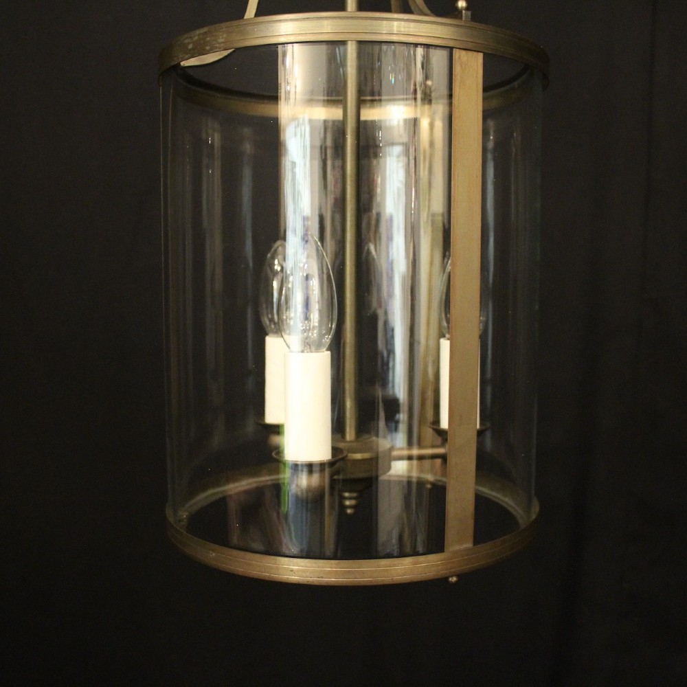 A cylindrical gilt brass hall lantern featuring curved clear glass panels, a central three-light candle fixture, and a suspended hanging chain. View III