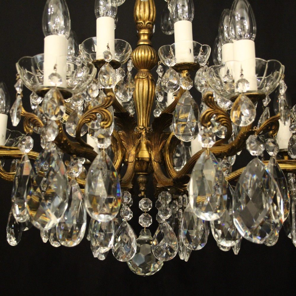 A multi-tiered antique French ceiling chandelier made of gilded brass with sixteen candle-style lights and faceted crystal drops against a dark background. View IV