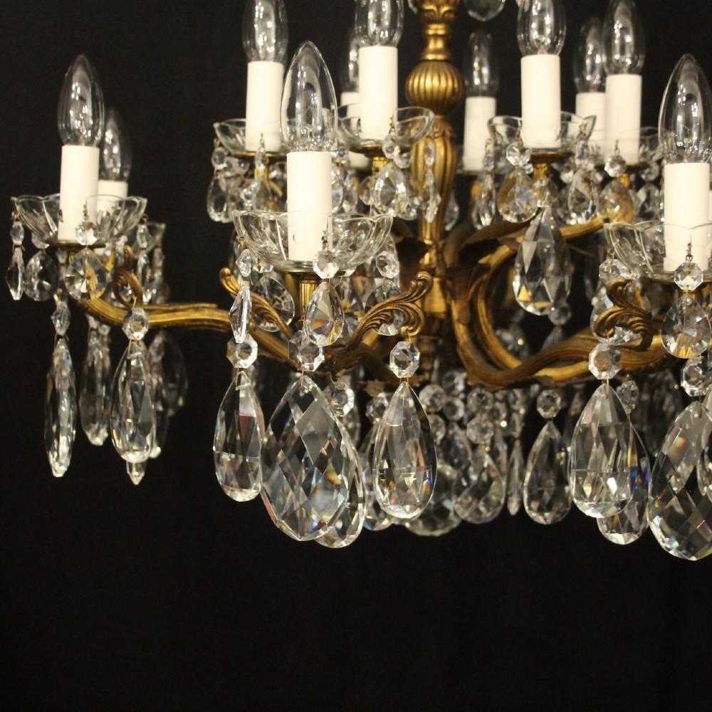 A multi-tiered antique French ceiling chandelier made of gilded brass with sixteen candle-style lights and faceted crystal drops against a dark background. View V