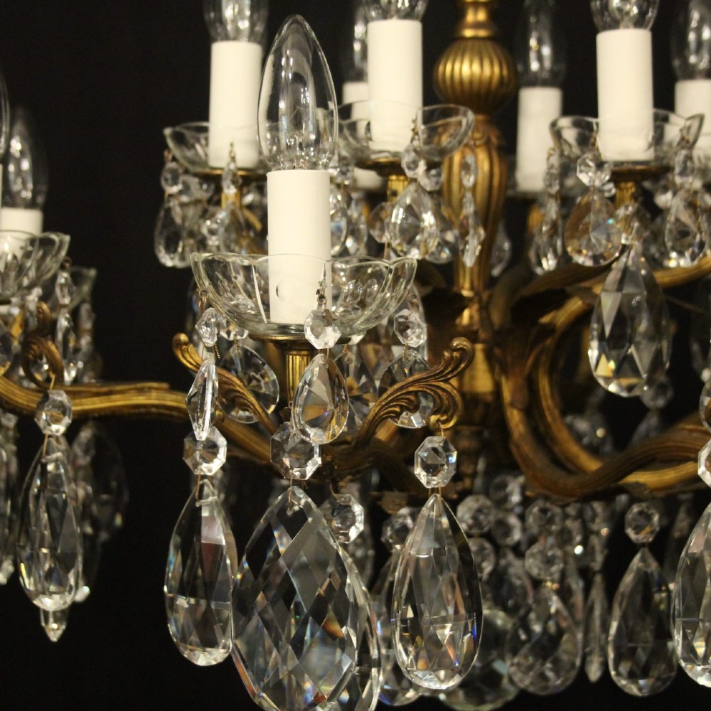A multi-tiered antique French ceiling chandelier made of gilded brass with sixteen candle-style lights and faceted crystal drops against a dark background. View VI