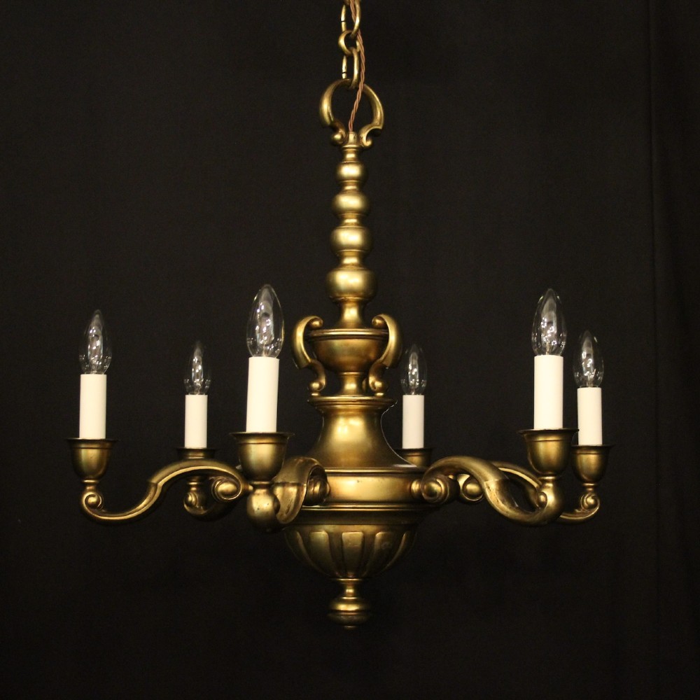 An antique warm brass chandelier featuring six curved scroll arms radiating from a central baluster column with white faux candle sockets.