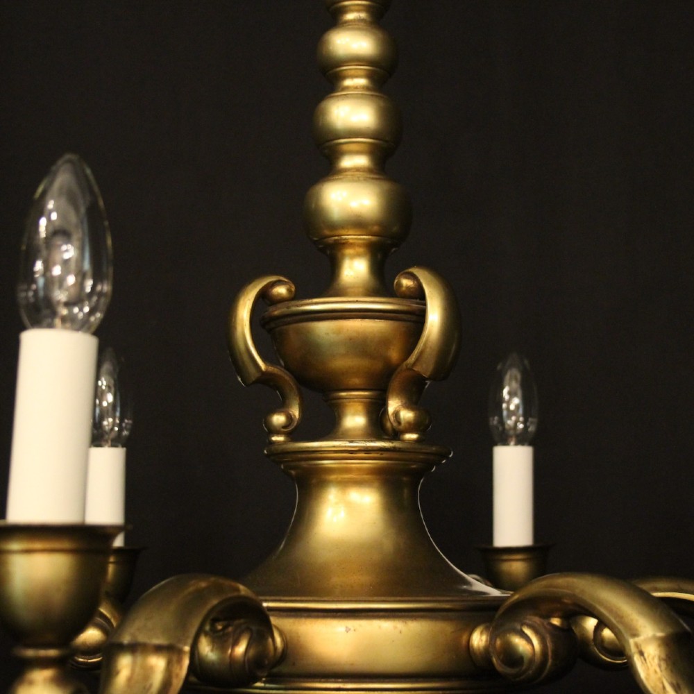 An antique warm brass chandelier featuring six curved scroll arms radiating from a central baluster column with white faux candle sockets. View III