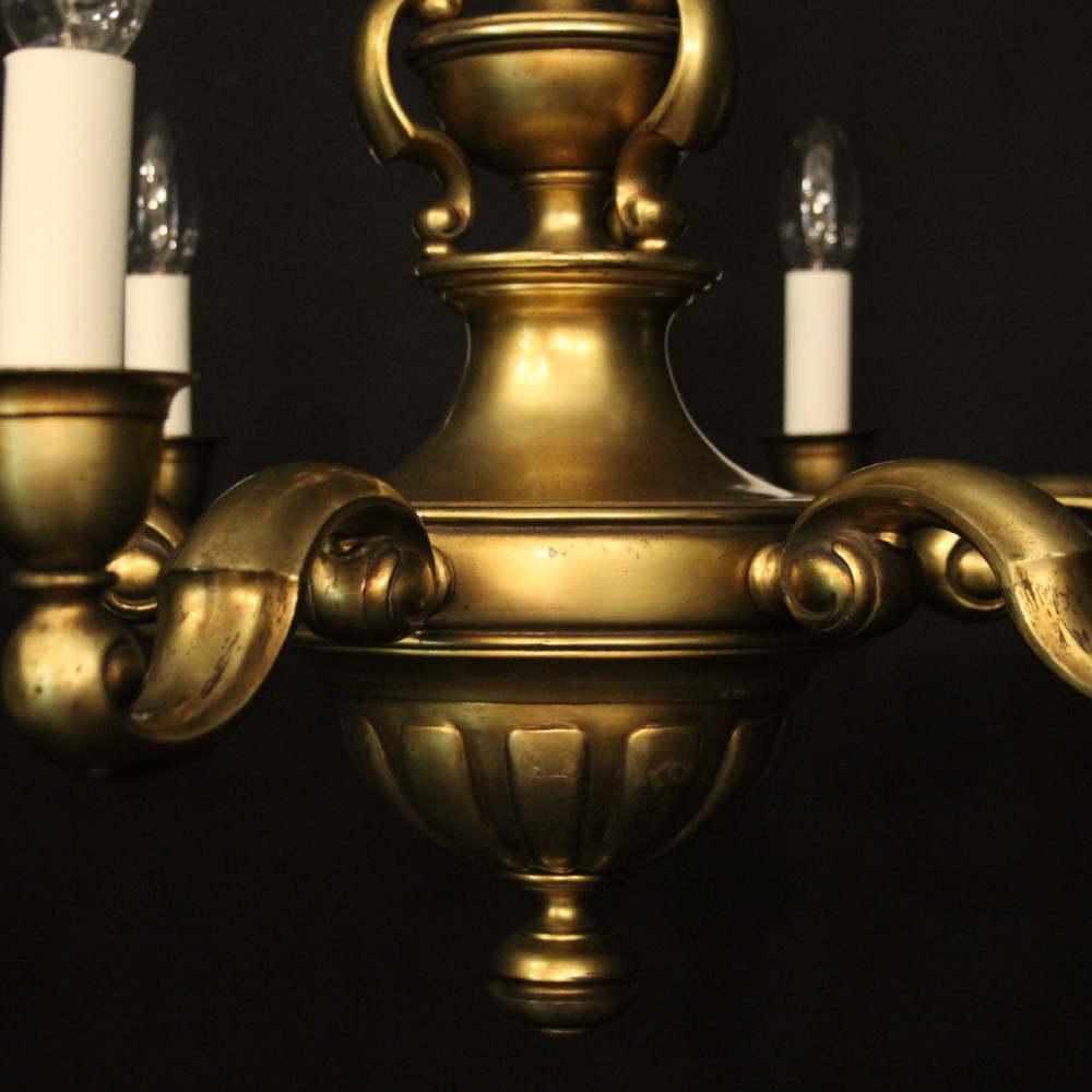 An antique warm brass chandelier featuring six curved scroll arms radiating from a central baluster column with white faux candle sockets. View IV