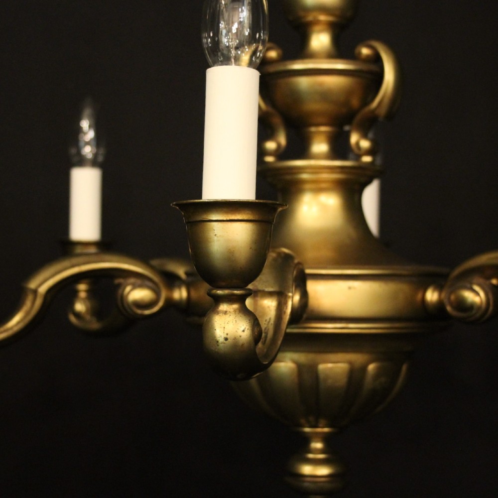 An antique warm brass chandelier featuring six curved scroll arms radiating from a central baluster column with white faux candle sockets. View V