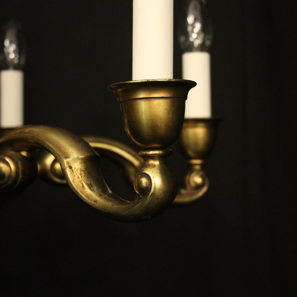 An antique warm brass chandelier featuring six curved scroll arms radiating from a central baluster column with white faux candle sockets. View VI
