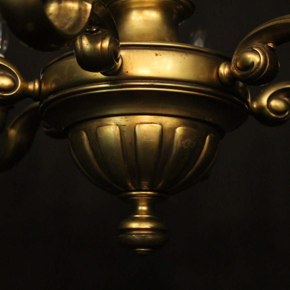 An antique warm brass chandelier featuring six curved scroll arms radiating from a central baluster column with white faux candle sockets. View VIII