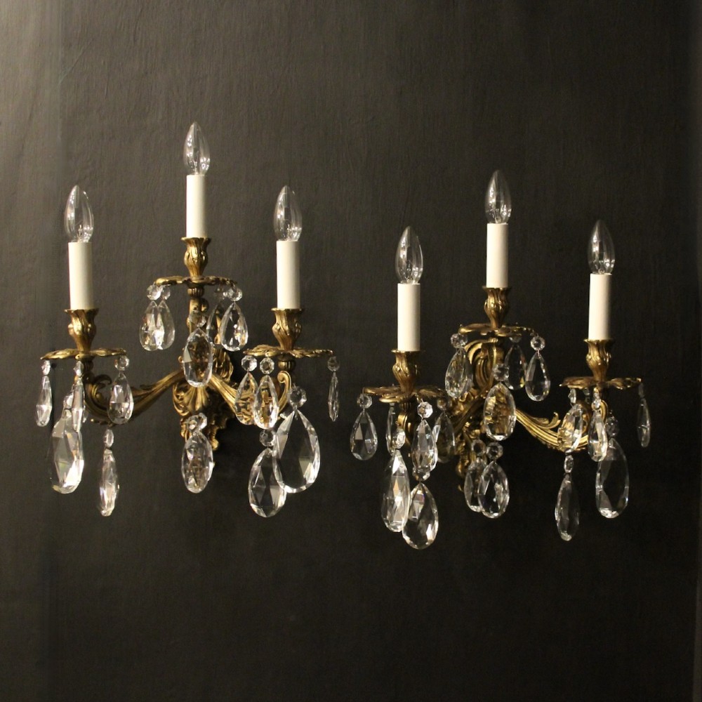 A pair of French three-arm gilded metal wall sconces adorned with hanging clear glass crystal droplets and white candle sleeves.