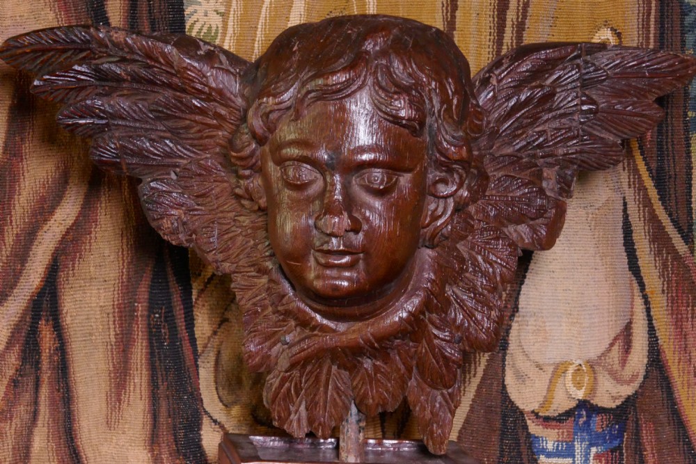 A dark carved oak winged cherub head with detailed feathering and curled hair, mounted upon a wooden square plinth base. View II