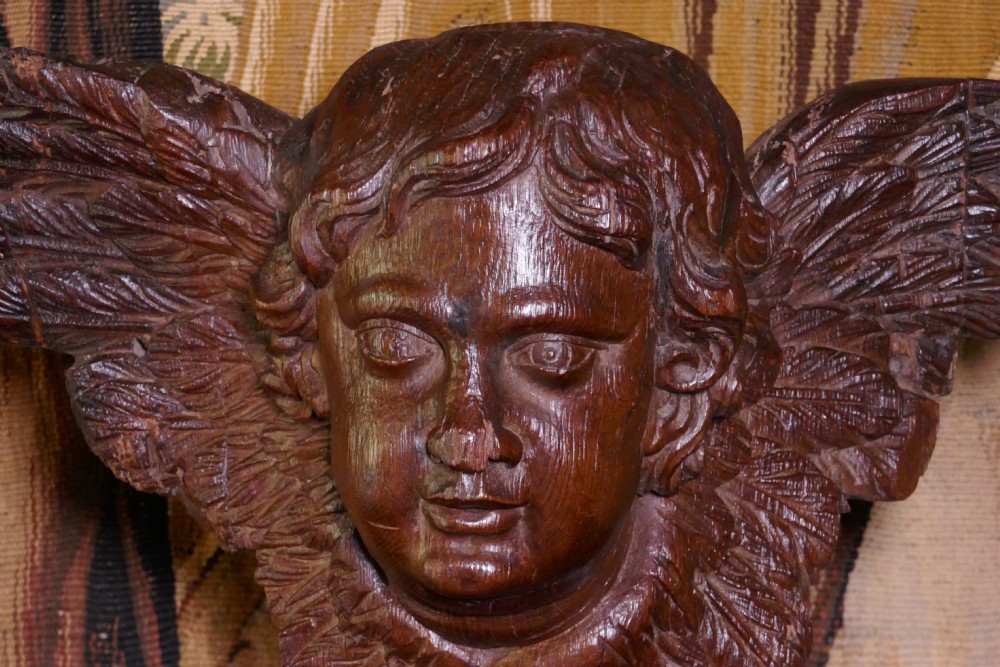 A dark carved oak winged cherub head with detailed feathering and curled hair, mounted upon a wooden square plinth base. View III