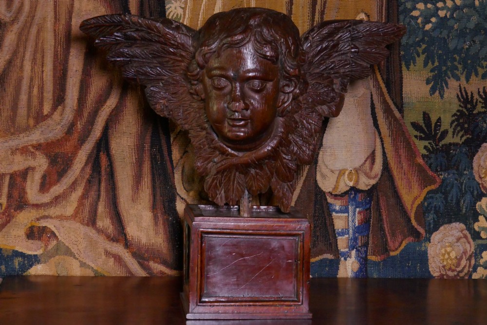 A dark carved oak winged cherub head with detailed feathering and curled hair, mounted upon a wooden square plinth base. View X