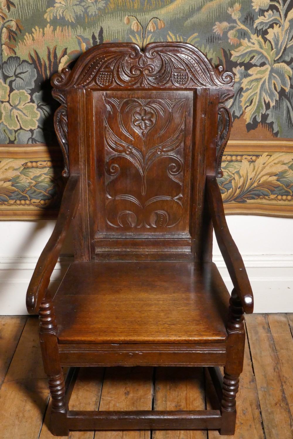 A 17th-century dark oak wainscot chair with a carved floral panel, arched cresting rail, and turned front legs.