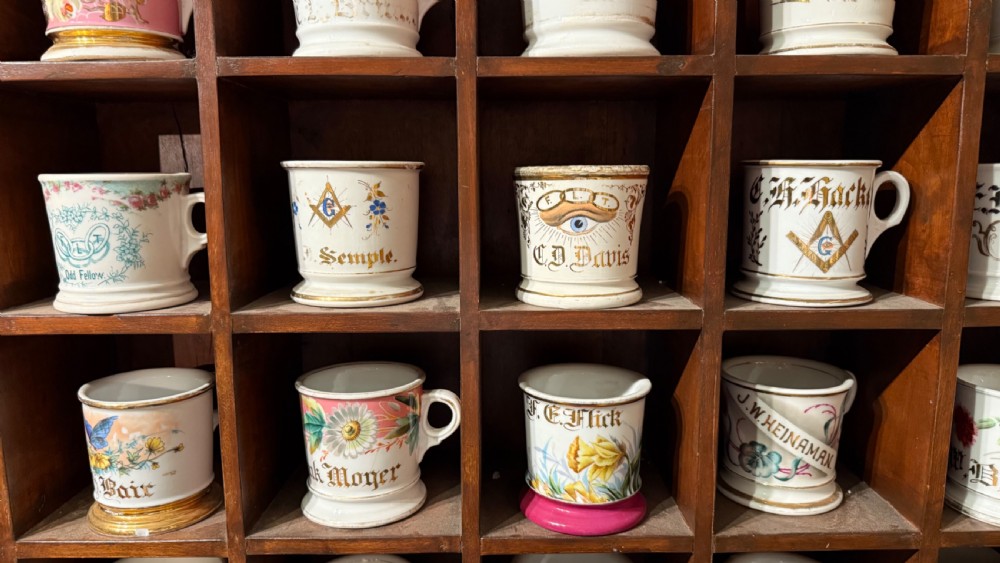 A dark mahogany cubby shelf unit filled with individual hand-painted ceramic shaving mugs featuring gilded names and motifs. View XIV