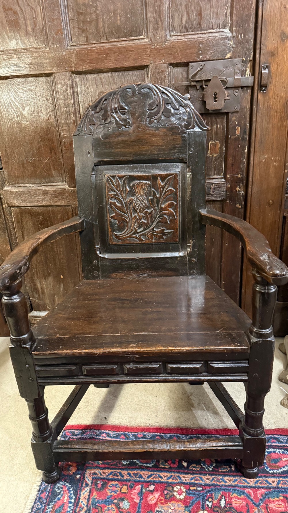 A 17th-century dark oak wainscot armchair with a carved thistle back panel, carved top crest, and turned legs. View V