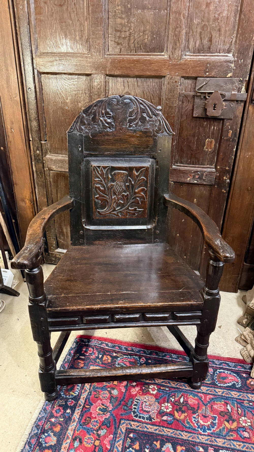 A 17th-century dark oak wainscot armchair with a carved thistle back panel, carved top crest, and turned legs.