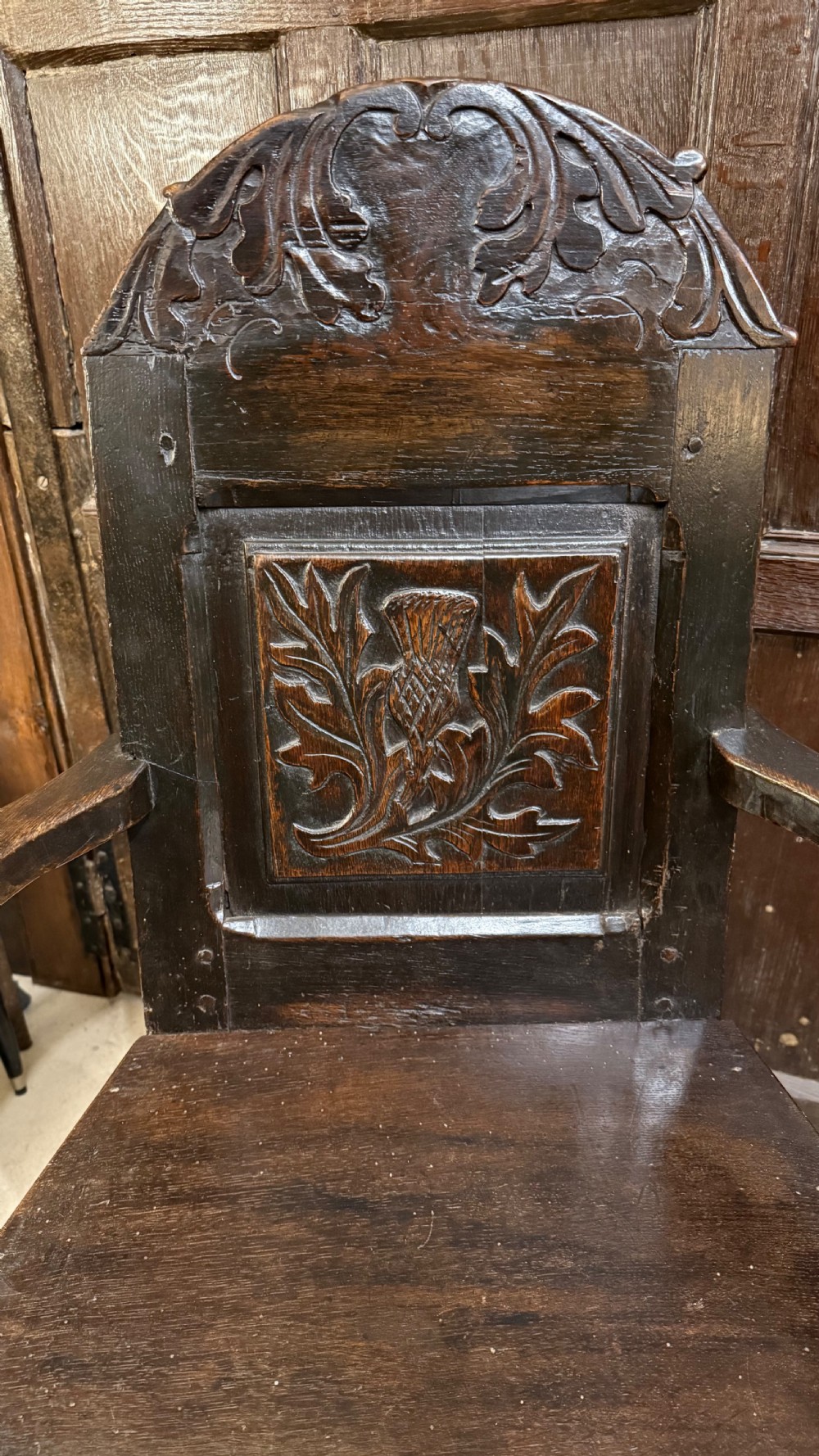 A 17th-century dark oak wainscot armchair with a carved thistle back panel, carved top crest, and turned legs. View X