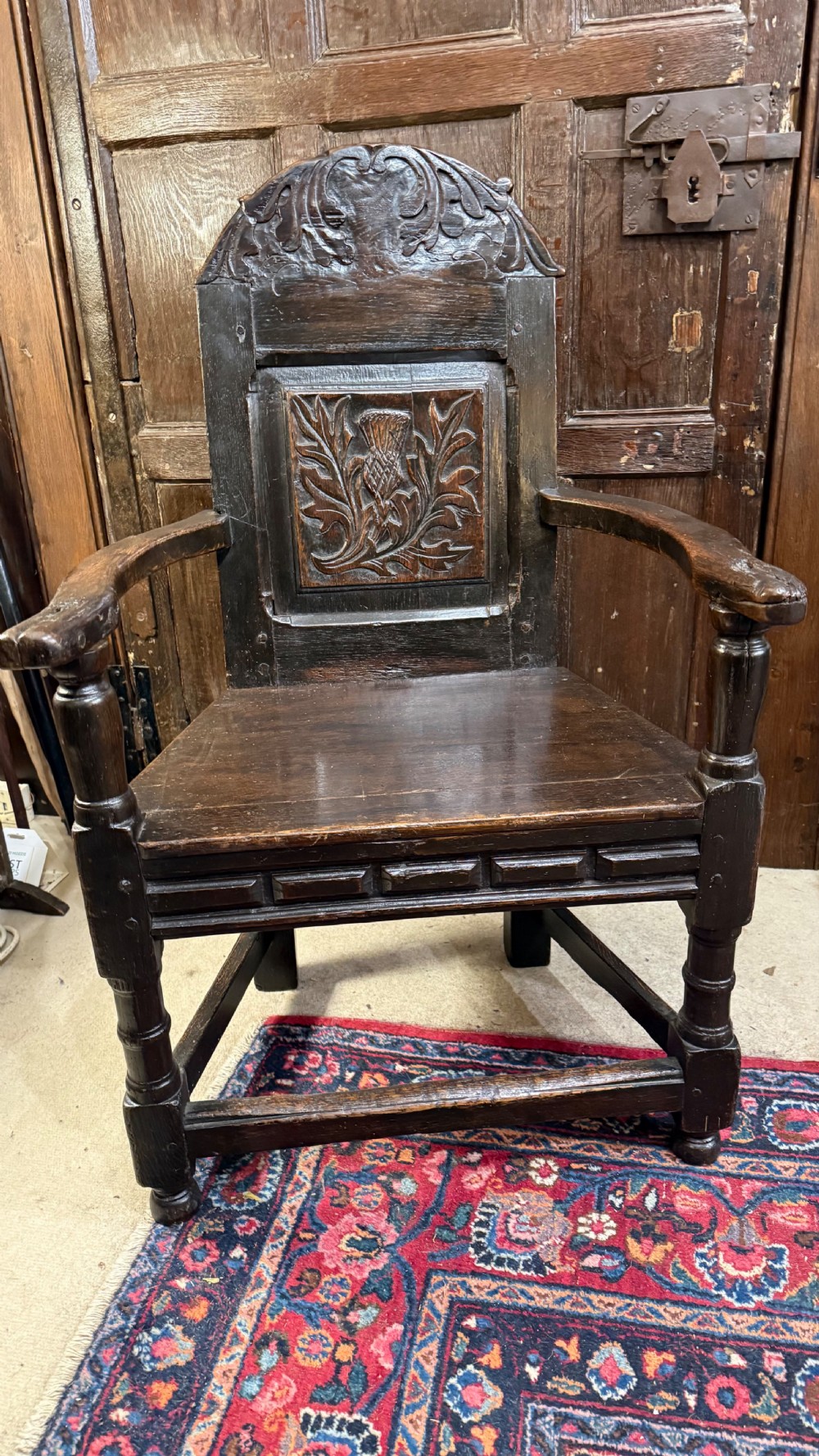 A 17th-century dark oak wainscot armchair with a carved thistle back panel, carved top crest, and turned legs. View XIV