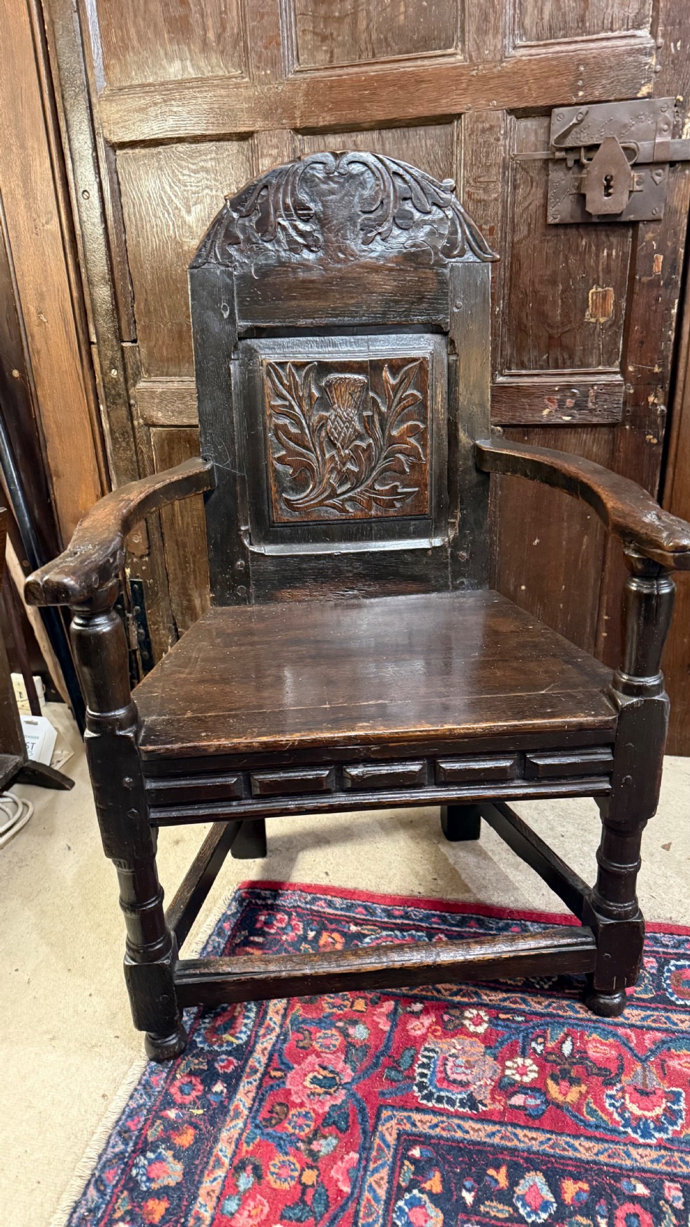 A 17th-century dark oak wainscot armchair with a carved thistle back panel, carved top crest, and turned legs. View XV