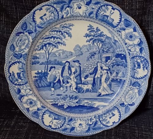 fig 1. A circular blue and white transfer-printed earthenware plate featuring a central pastoral scene of figures near beehives surrounded by a floral and animal vignette border.