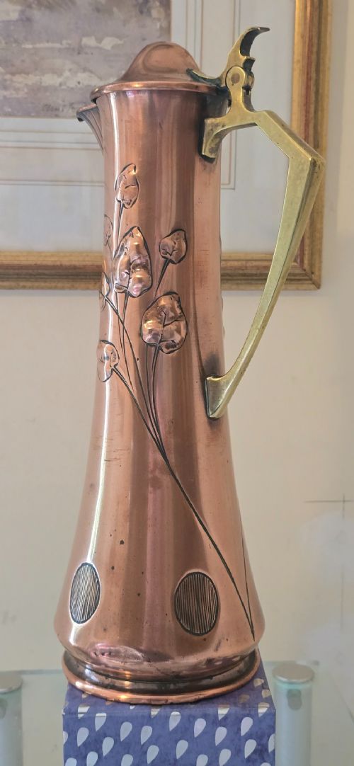 fig 4. An Art Nouveau tapered copper ewer with a raised floral relief pattern, angular brass handle, hinged brass cap, and subtle dark circular motifs near the base.