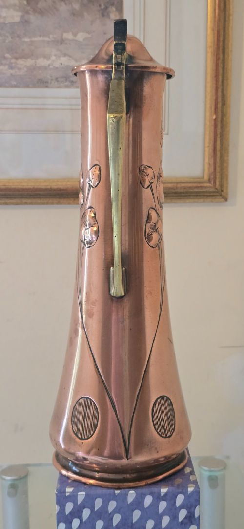 fig 5. An Art Nouveau tapered copper ewer with a raised floral relief pattern, angular brass handle, hinged brass cap, and subtle dark circular motifs near the base.