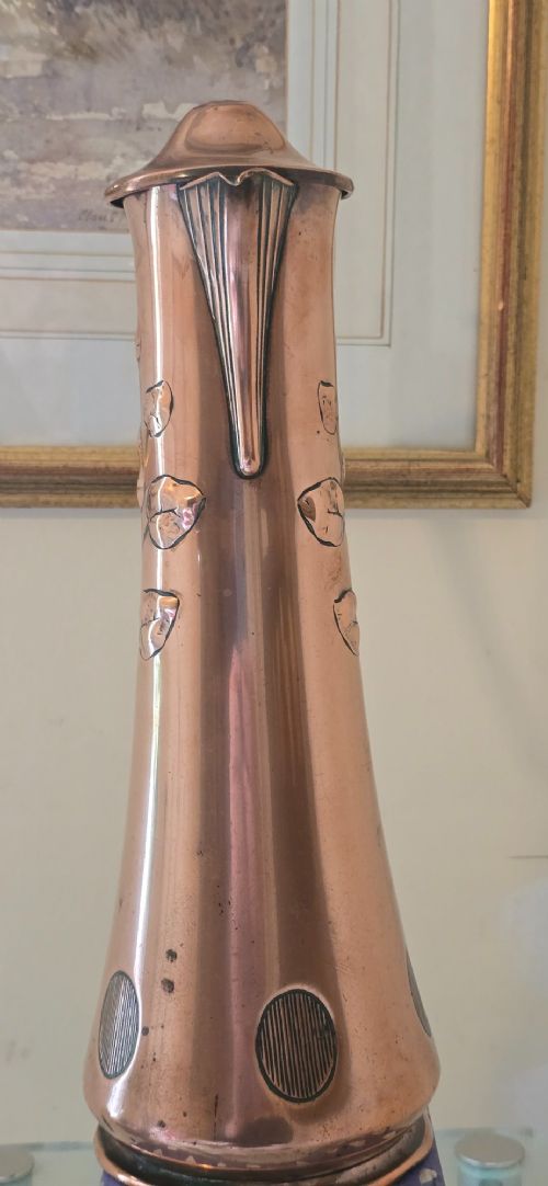 fig 6. An Art Nouveau tapered copper ewer with a raised floral relief pattern, angular brass handle, hinged brass cap, and subtle dark circular motifs near the base.