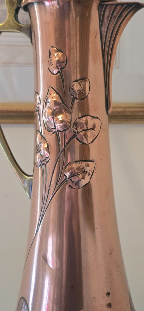 fig 7. An Art Nouveau tapered copper ewer with a raised floral relief pattern, angular brass handle, hinged brass cap, and subtle dark circular motifs near the base.