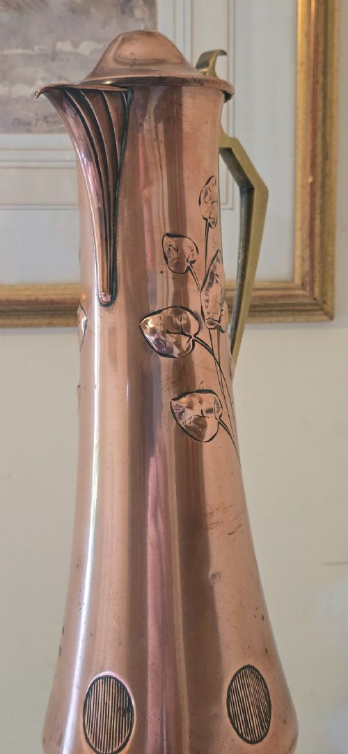 fig 8. An Art Nouveau tapered copper ewer with a raised floral relief pattern, angular brass handle, hinged brass cap, and subtle dark circular motifs near the base.