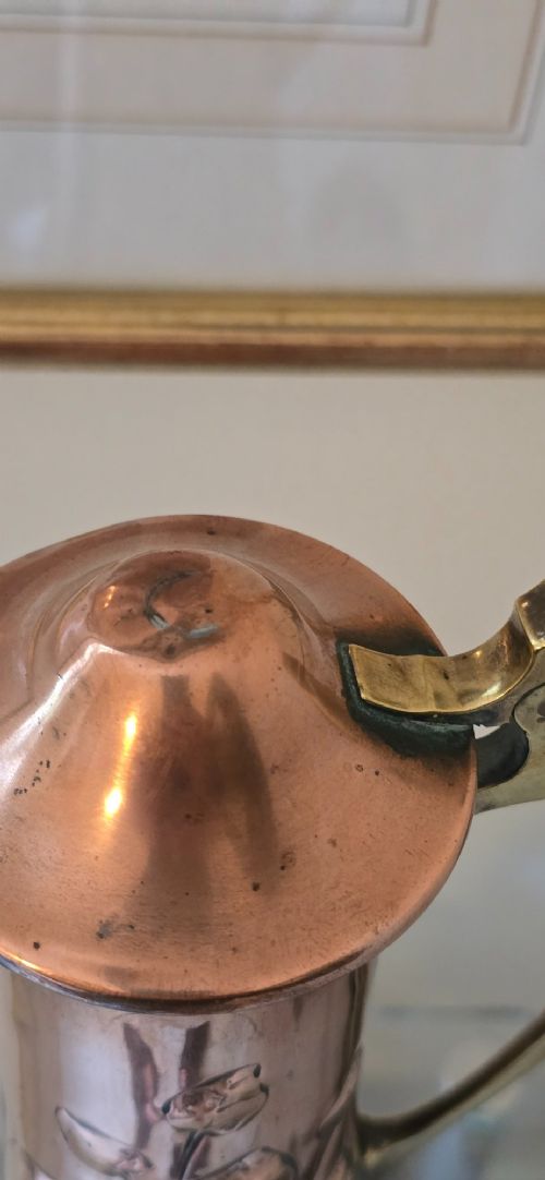 An Art Nouveau tapered copper ewer with a raised floral relief pattern, angular brass handle, hinged brass cap, and subtle dark circular motifs near the base. View XI