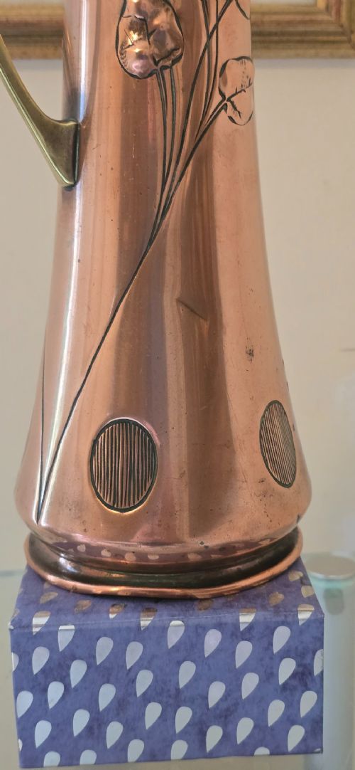 An Art Nouveau tapered copper ewer with a raised floral relief pattern, angular brass handle, hinged brass cap, and subtle dark circular motifs near the base. View XII