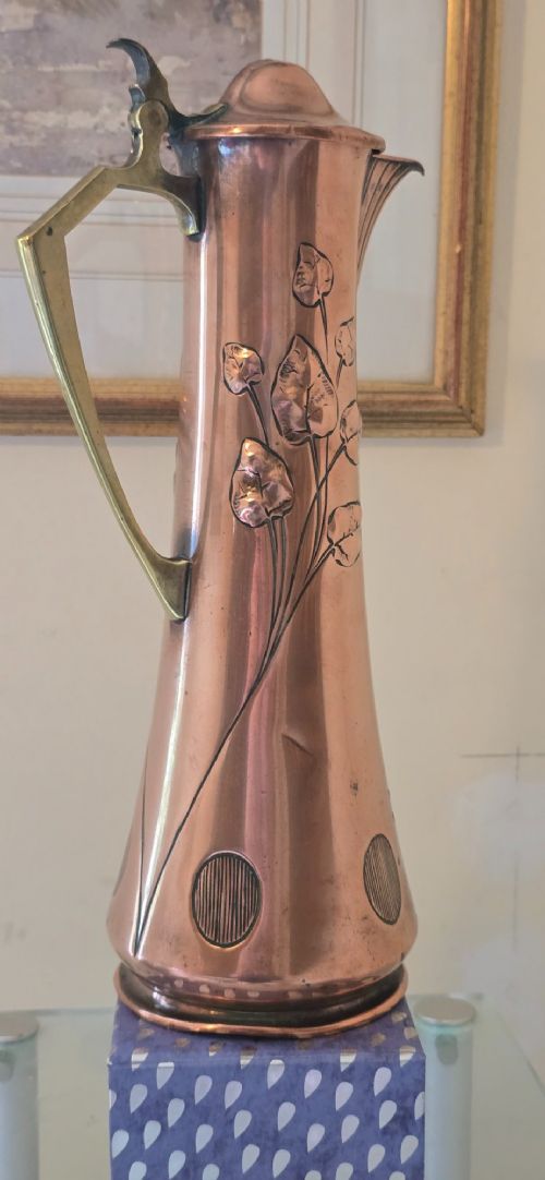 An Art Nouveau tapered copper ewer with a raised floral relief pattern, angular brass handle, hinged brass cap, and subtle dark circular motifs near the base. View XIII