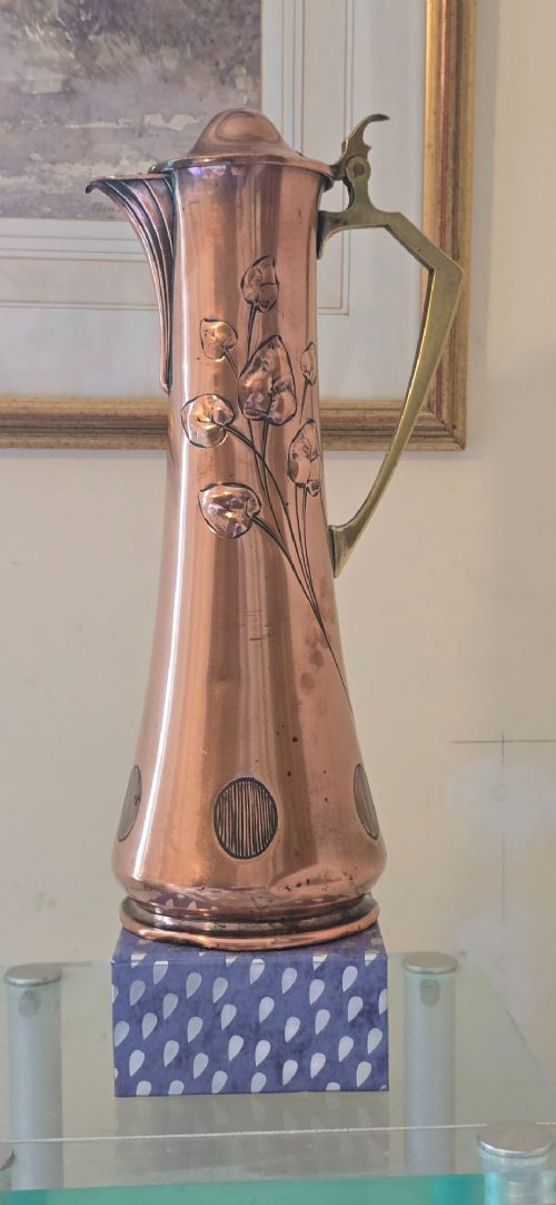 An Art Nouveau tapered copper ewer with a raised floral relief pattern, angular brass handle, hinged brass cap, and subtle dark circular motifs near the base. View XIV