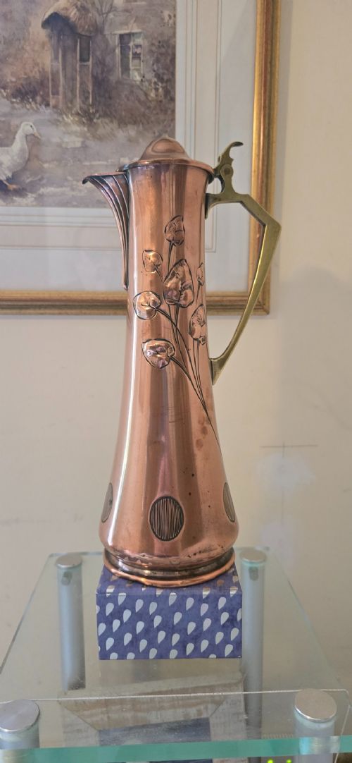 An Art Nouveau tapered copper ewer with a raised floral relief pattern, angular brass handle, hinged brass cap, and subtle dark circular motifs near the base. View XV