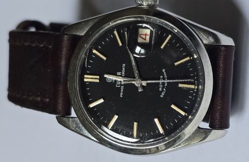 fig 2. A vintage Tudor Prince Oysterdate wristwatch with a stainless steel case, a black dial featuring baton hour markers, a date aperture at three oclock, and a dark brown leather strap.