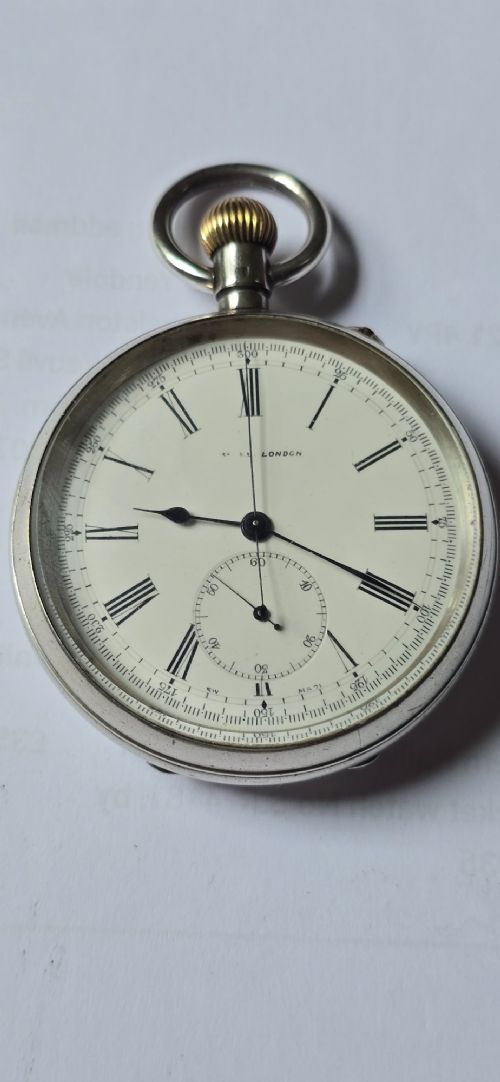 fig 1. An open-face silver pocket watch featuring a white enamel dial with black Roman numerals, split-seconds hands, a subsidiary seconds dial, and a brass onion crown at the top.