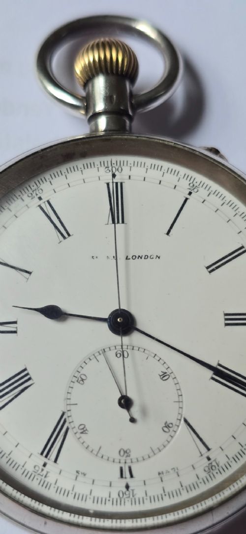 fig 2. An open-face silver pocket watch featuring a white enamel dial with black Roman numerals, split-seconds hands, a subsidiary seconds dial, and a brass onion crown at the top.