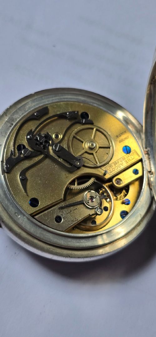 fig 5. An open-face silver pocket watch featuring a white enamel dial with black Roman numerals, split-seconds hands, a subsidiary seconds dial, and a brass onion crown at the top.
