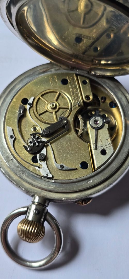 fig 6. An open-face silver pocket watch featuring a white enamel dial with black Roman numerals, split-seconds hands, a subsidiary seconds dial, and a brass onion crown at the top.
