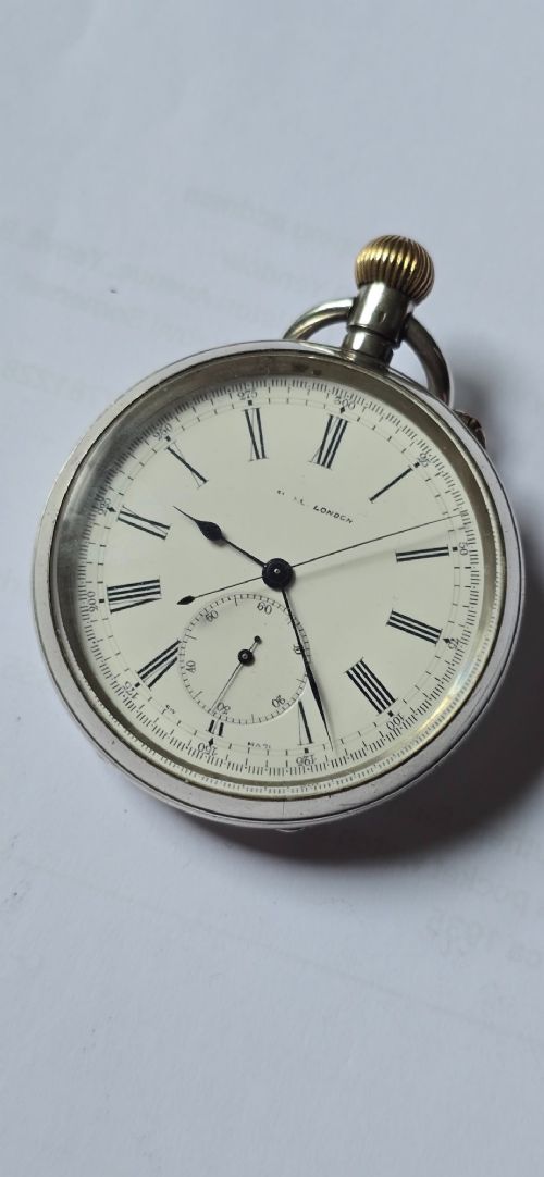 An open-face silver pocket watch featuring a white enamel dial with black Roman numerals, split-seconds hands, a subsidiary seconds dial, and a brass onion crown at the top. View X