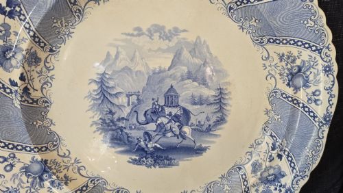 fig 2. An antique circular earthenware plate featuring a blue transfer-printed central scene of Hannibal with an elephant and figures in a mountainous landscape, surrounded by an ornate scalloped floral border.