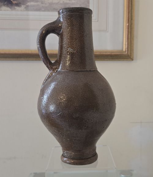 fig 1. A 17th-century dark brown salt-glazed stoneware bottle featuring a bulbous body, narrow neck, turned base, and a single loop handle.