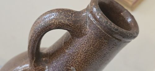 fig 4. A 17th-century dark brown salt-glazed stoneware bottle featuring a bulbous body, narrow neck, turned base, and a single loop handle.